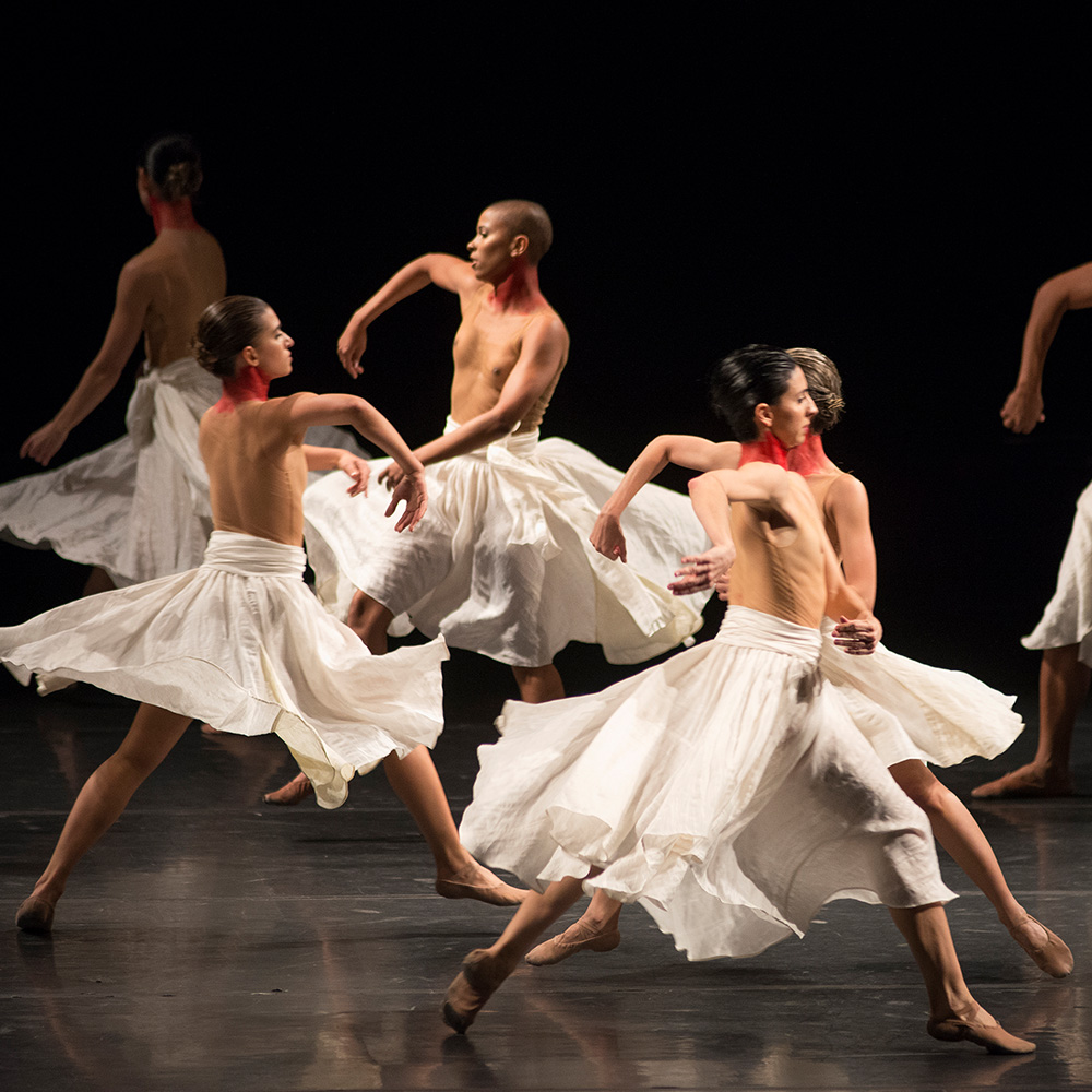 Algumas bailarinas do Grupo Corpo estão em um movimento de dança, vestindo uma saia branca.