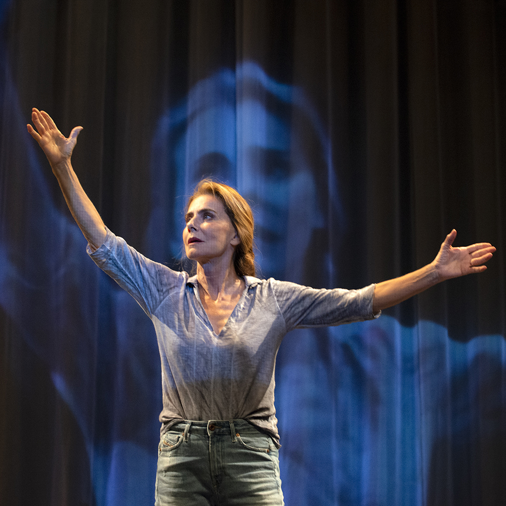 A imagem mostra a atriz Maitê Proença está de braços abertos, olhando para cima. Ela veste uma blusa cinza e o fundo da imagem é azul, com uma foto dela refletida.
