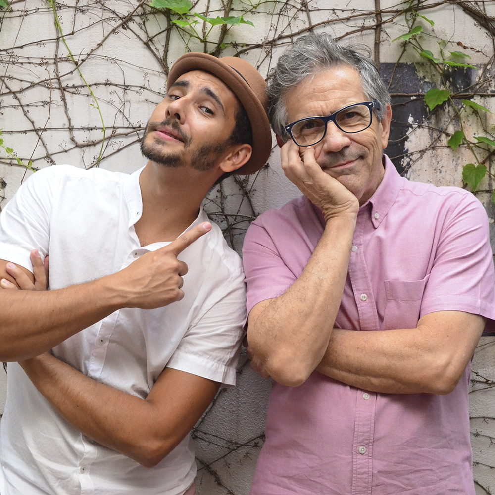 A foto do grupo Siricutico mostra dois músicos um ao lado do outro. O músico da esquerda está com o dedo indicador apontado para o músico da direita, veste camisa branca, além de um chapéu marrom. O músico da direita está com uma das mãos apoiando o queixo e veste camisa rosa, além de usar óculos.