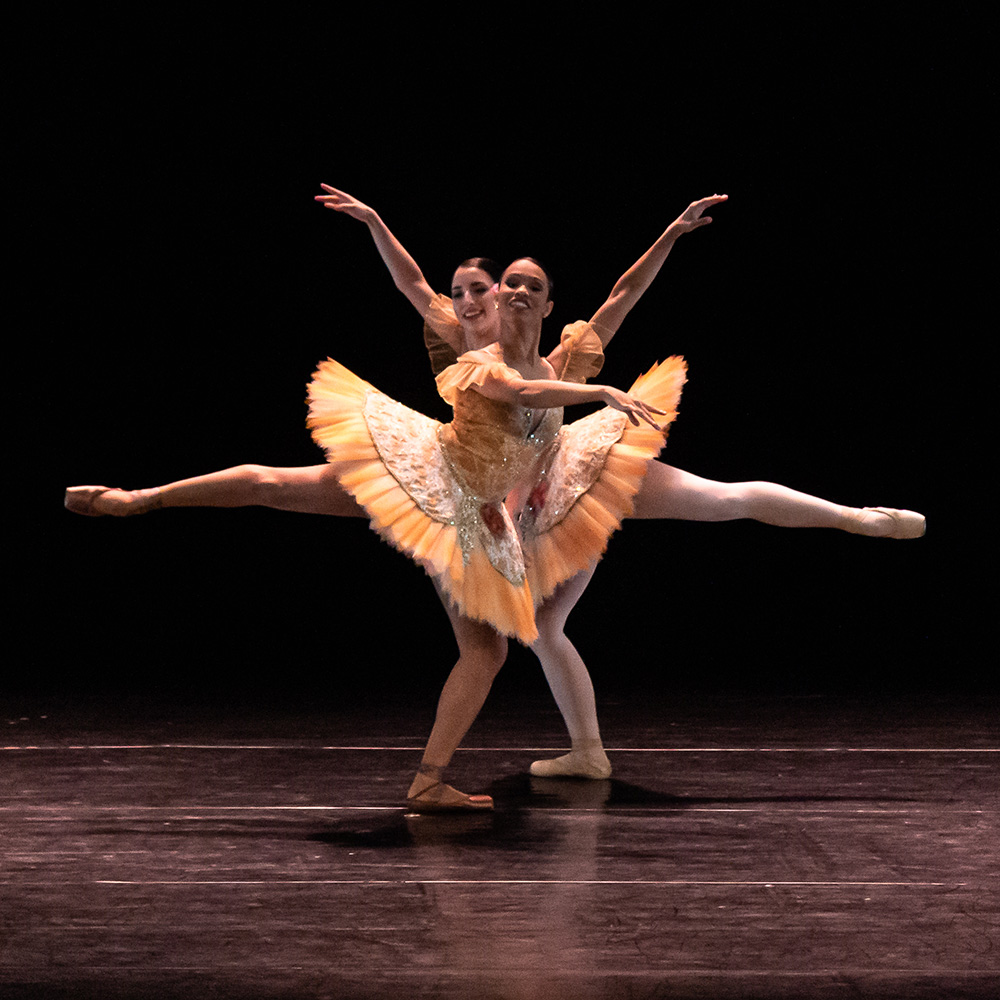 A foto mostra duas bailarinas com roupas em tons marrons, ambas em movimento de dança.