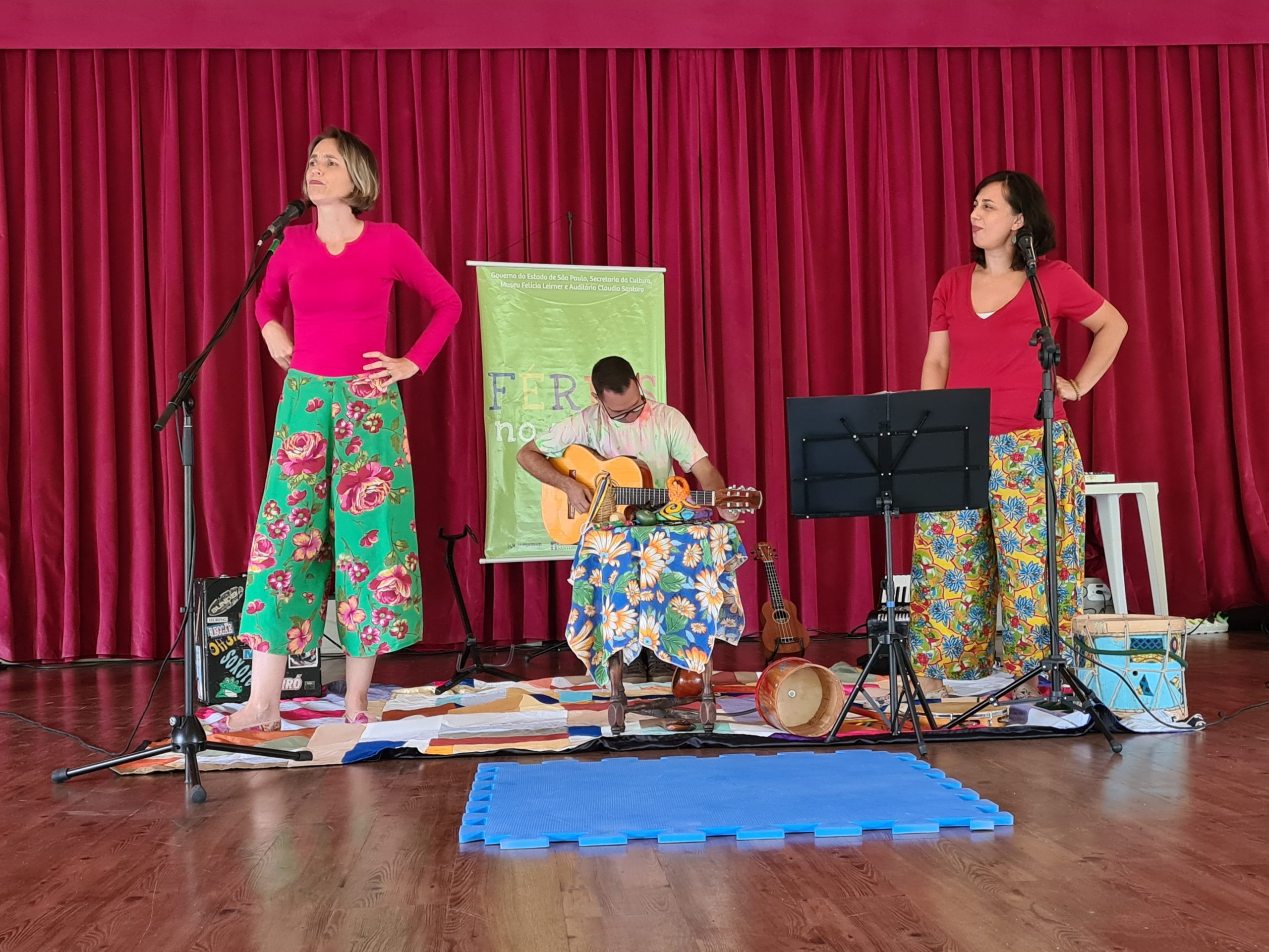 A imagem mostra uma dupla de contadoras de história acompanhada de um violonista. Elas estão usando camiseta rosa e calça florida. Ao redor há alguns elementos musicais como: acordeon, ukulele e tambores.