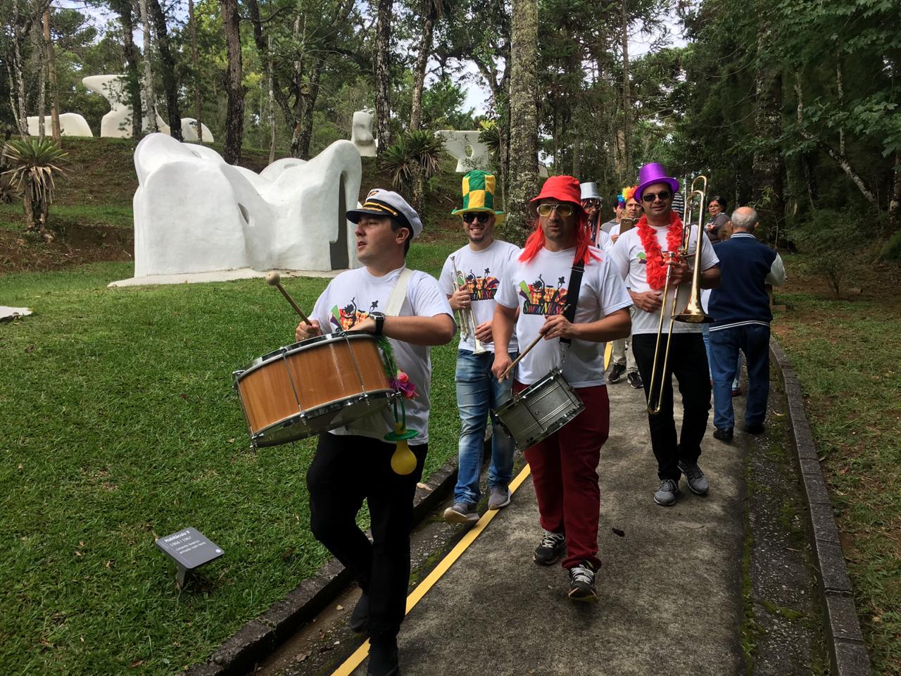 Na imagem, uma banda está caminhando pelas alamedas do Museu Felícia Leirner tocando instrumentos de sopro e percussão. Os integrantes da banda estão utilizando alguns objetos carnavalescos.