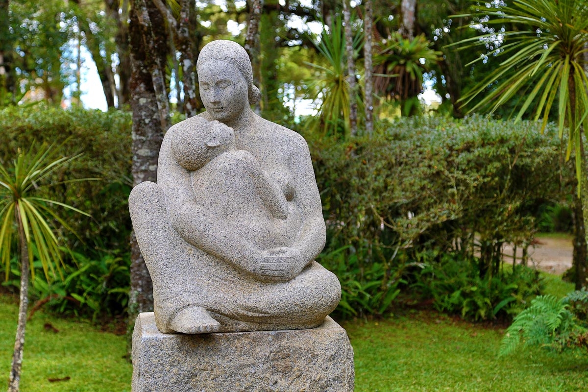Escultura figurativa feita em granito que representa uma mãe sentada segurando seu filho nos braços.