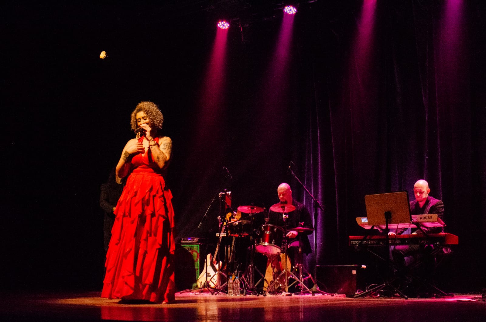 Na imagem a cantora Lia de Oliveira está cantando em um palco com luzes rosa. Ela está utilizando um vestido vermelho longo. Ao fundo há um homem tocando bateria e outro tocando teclado.