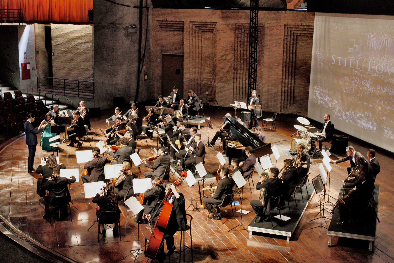 A imagem mostra um maestro regendo uma orquestra composta por diversos tipos de instrumentos.