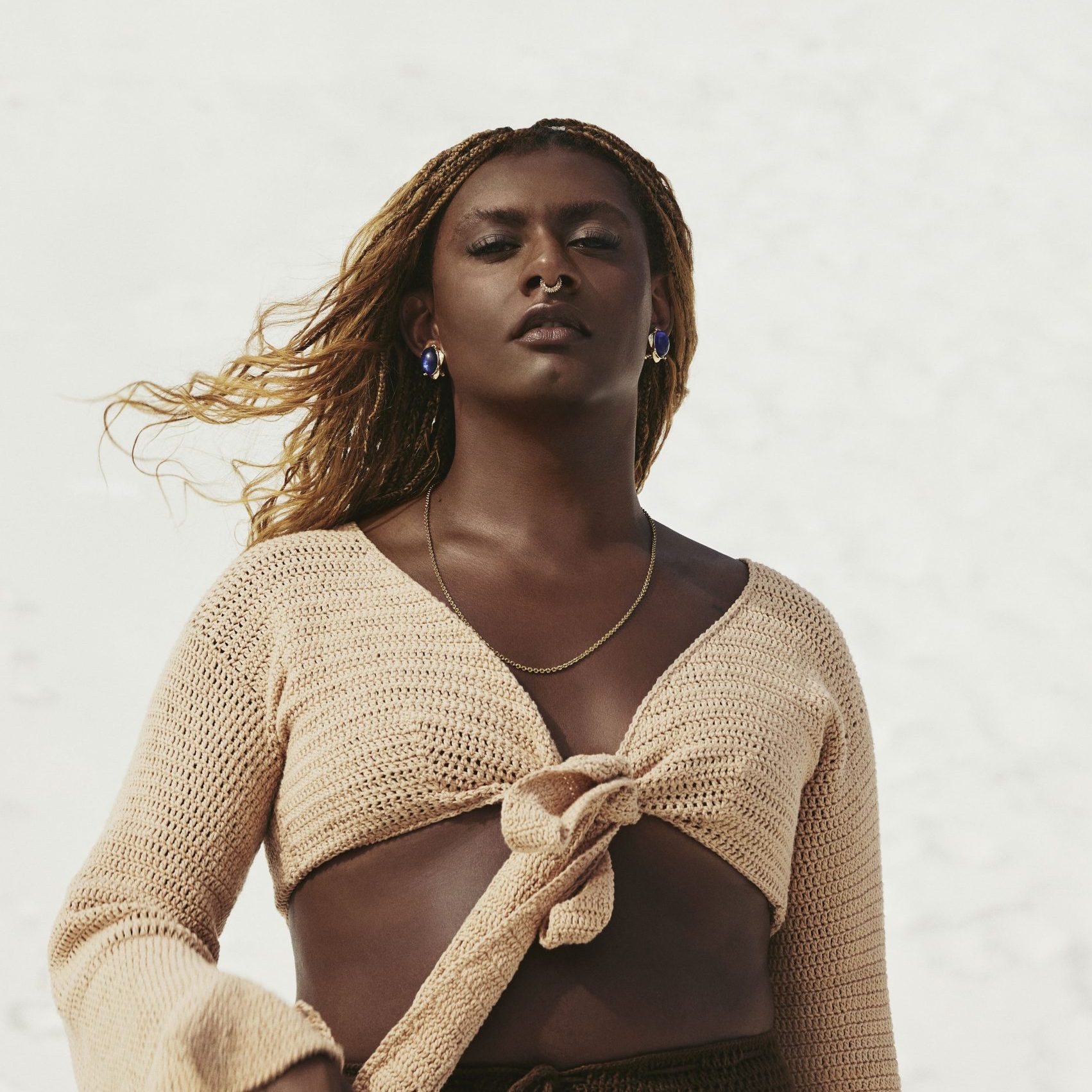 A imagem mostra a cantora Liniker. Ela é uma mulher preta, com os cabelos loiros nas pontas, que veste um cropped cor de areia.