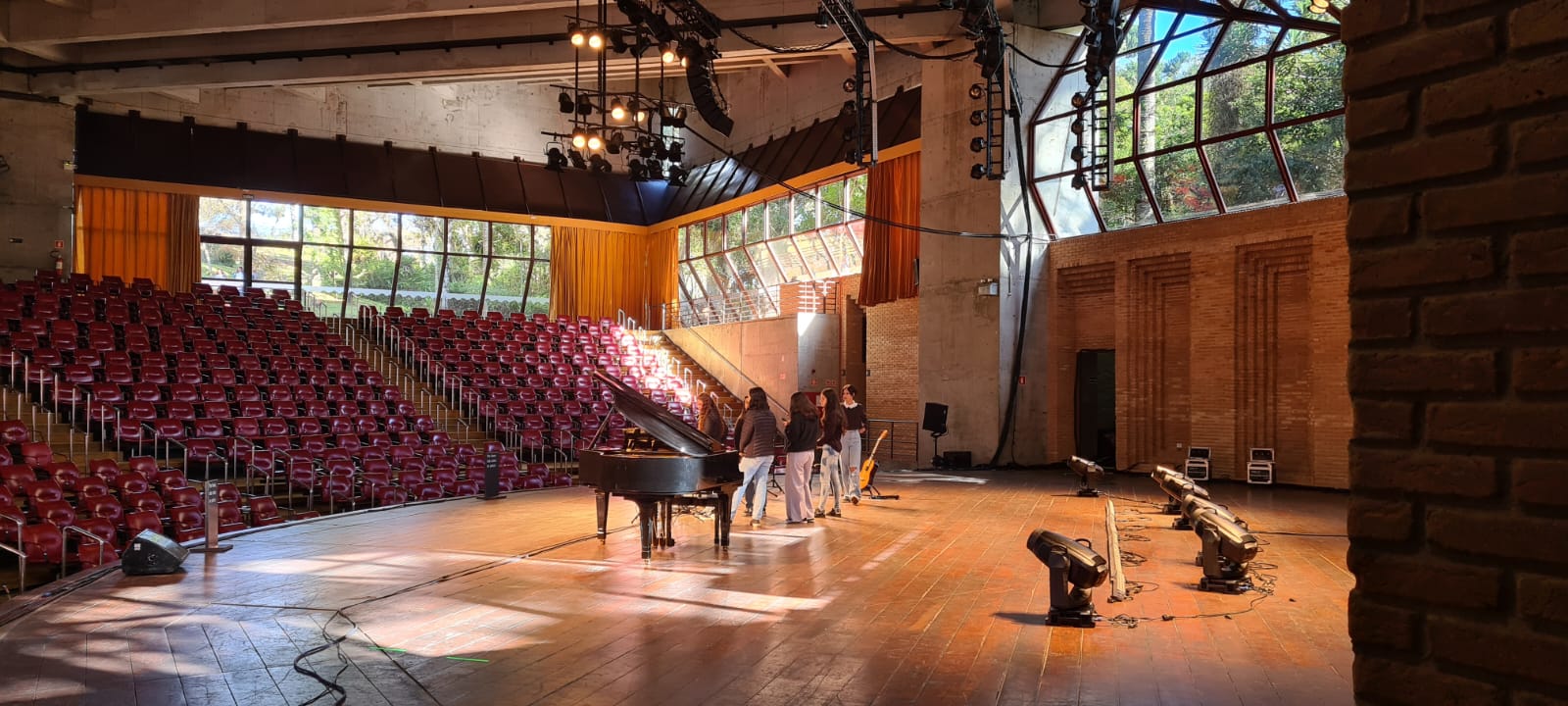 Na imagem, um grupo de jovens está no palco do Auditório Claudio Santoro se apresentando. No palco há um piano de calda e um violão.