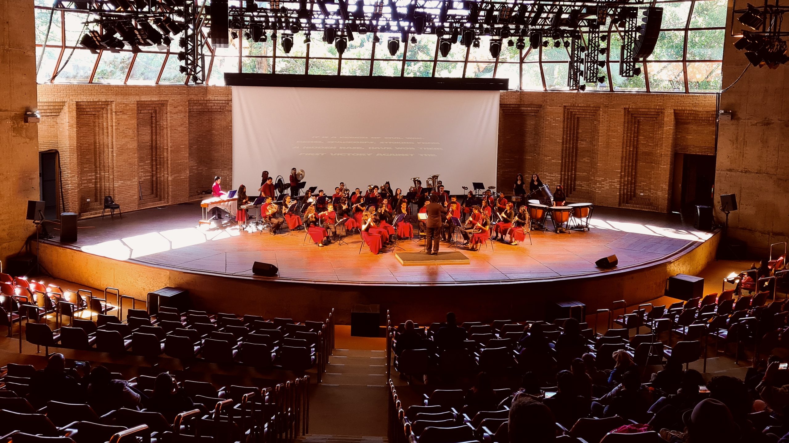 Crianças e adolescentes sentados em um palco tocam diferentes tipos de instrumentos, compondo uma orquestra. No centro, há uma homem de preto que faz a regência das crianças.