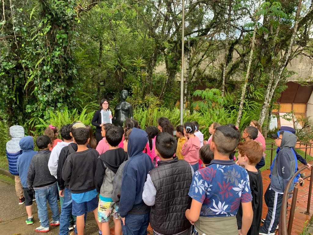 Na imagem um grupo de crianças estão prestando atenção na educadora que segura em suas mãos uma placa com a imagem da artista Felícia Leirner. Eles estão ao ar livre, próximos de uma escultura feita em bronze.