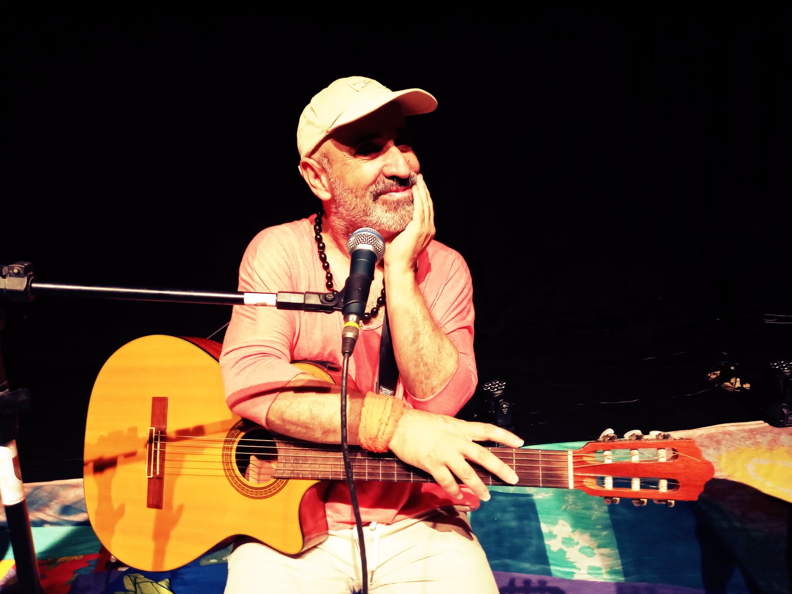 Nando luz é um homem branco que está usando um boné de cor clara, uma camisa rosa e segura em seu colo um violão.