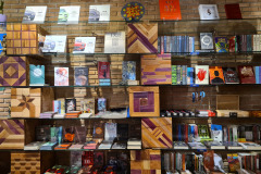 Na imagem, há prateleiras de vidro e caixas de madeiras decorativas empilhadas formando uma estante de livros com diversos exemplares de gêneros como fantasia, romance, terror, literatura nacional.