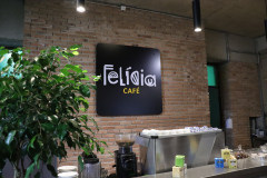 Na imagem há um balcão com uma maquina de fazer café. Ao fundo, uma parede de tijolos com uma placa com o logo "Felícia Café".