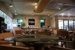 Na imagem há mesas e cadeiras dispostas no espaço do restaurante e café, no qual, ao fundo há uma parede de tijolos com uma placa com a logo "Felícia Café".