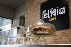 Na imagem há uma boleira de vidro com um bolo. Atrás há uma parede de tijolos com uma placa com o logo "Felícia Café"