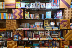 Na imagem, há prateleiras de vidro e caixas de madeiras decorativas empilhadas formando uma estante de livros com diversos exemplares de livros infantis.