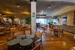 Foto da área integrada entre restaurante Felícia Café e a Livraria História Sem Fim. Na imagem há mesas e cadeiras dispostas em um espaço onde, ao fundo, à esquerda, há uma estante de livros feita com prateleiras de vidro e caixas decorativas. À direita, há uma balcão com vitrine de salgados, boleiras e caixa de atendimento.